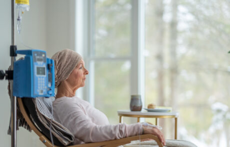 A senior woman sits in the comfort of her own home as she receives her chemotherapy treatment. She is dressed comfortably and wearing a headscarf to keep her warm.