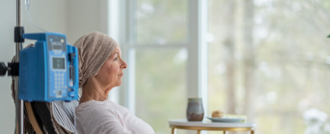 A senior woman sits in the comfort of her own home as she receives her chemotherapy treatment. She is dressed comfortably and wearing a headscarf to keep her warm.